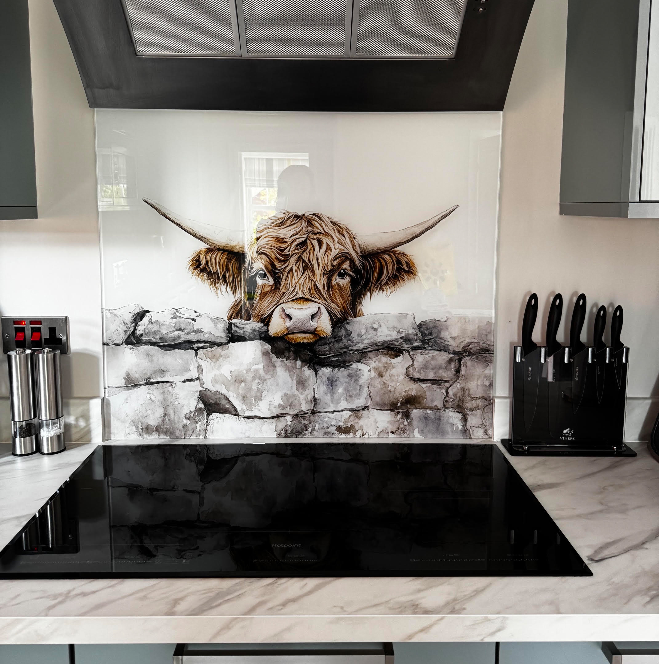 Highland cow printed glass splashback behind a black kitchen hob, featuring a Highland cow looking over a rustic stone wall in Gary and Ann’s modern grey and white kitchen.