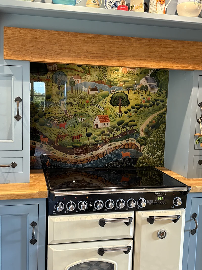 Folk Farmyard printed glass splashback installed behind a cream Rangemaster cooker in Rowena’s country-style kitchen with blue cabinets and wooden worktops.