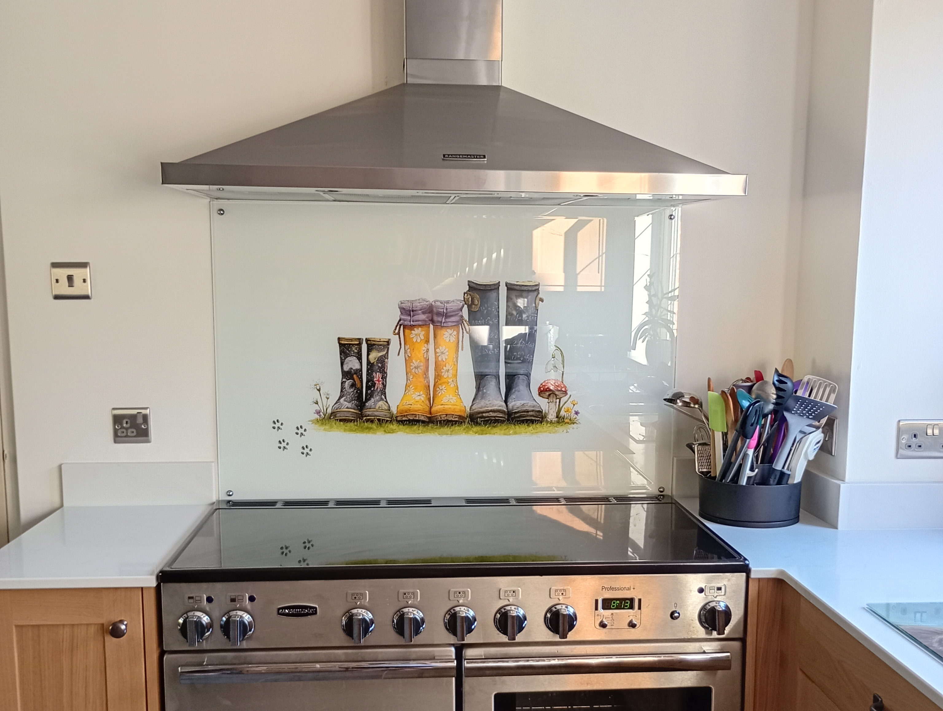 Large personalised family glass splashback behind a Rangemaster cooker featuring three pairs of Wellington boots inspired by flowers, maths, history and space, with dog paw prints beside them.